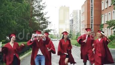 令人兴奋的毕业学生在校园里沿路奔跑，拿着毕业证书，穿着毕业礼服和帽子